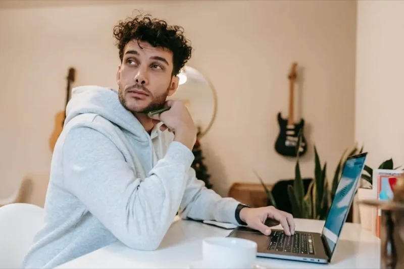 Freelancer trabalhando com notebook à sua frente, olhando em direção oposta com semblante pensativo.