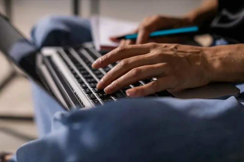 Pessoa com notebook no colo, mão esquerda no teclado, mão direita segurança um lápis em cima de um caderno apoiado em sua coxa.